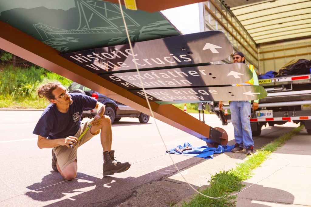 a man is kneeling on the ground underneath a large wayfinding sign. He is installing the navigational signs on the side of the road.