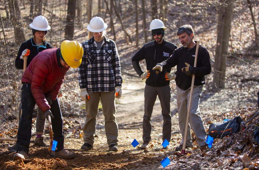 a group of five trail advocates gather together to discuss an upcoming project. they are all wearing work clothes and four of them have hard hats on. one of them is gesturing towards the trail.