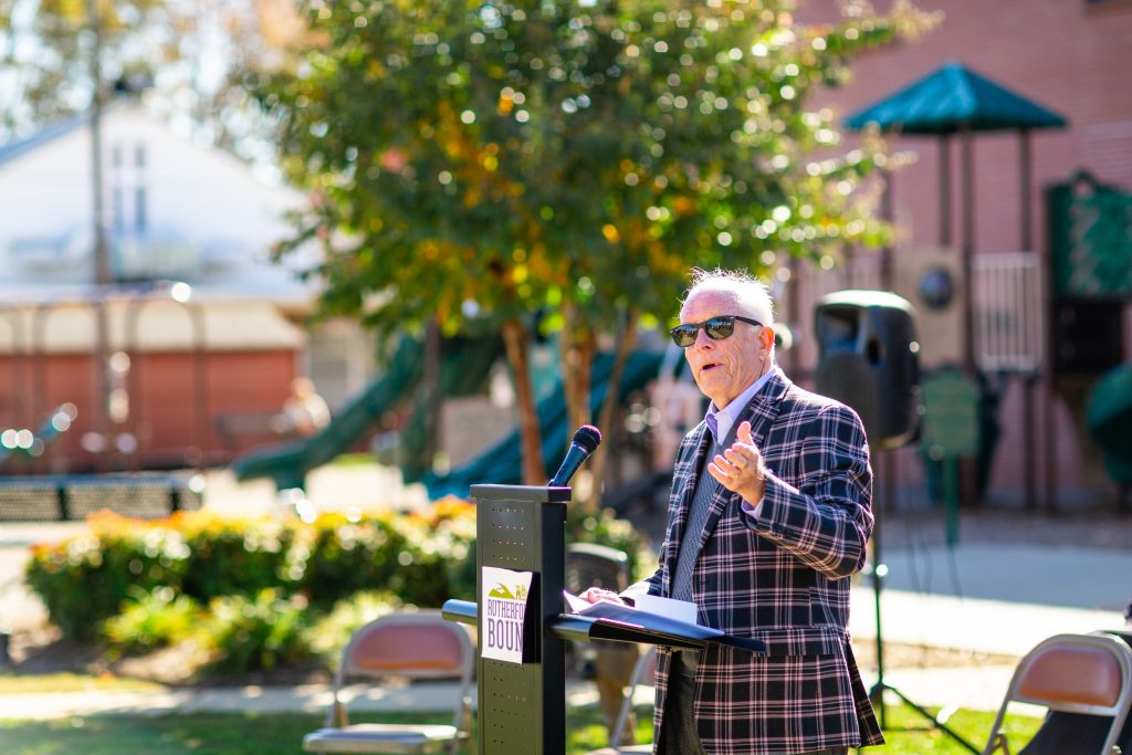 The mayor speaks to the crowd gathered at the ribbon cutting ceremony. He is wearing sunglasses and a purple plaid jacket.